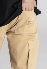 Beżowe spodnie cargo wykonane z wytrzymałej bawełny, z boczną kieszenią i subtelnym detalem logo. Materiał ma gładką fakturę i luźny krój.