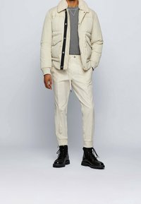 Giacca imbottita beige chiaro con colletto in shearling, maglietta grigia a maniche lunghe, pantaloni jogger panna e stivali neri con lacci.