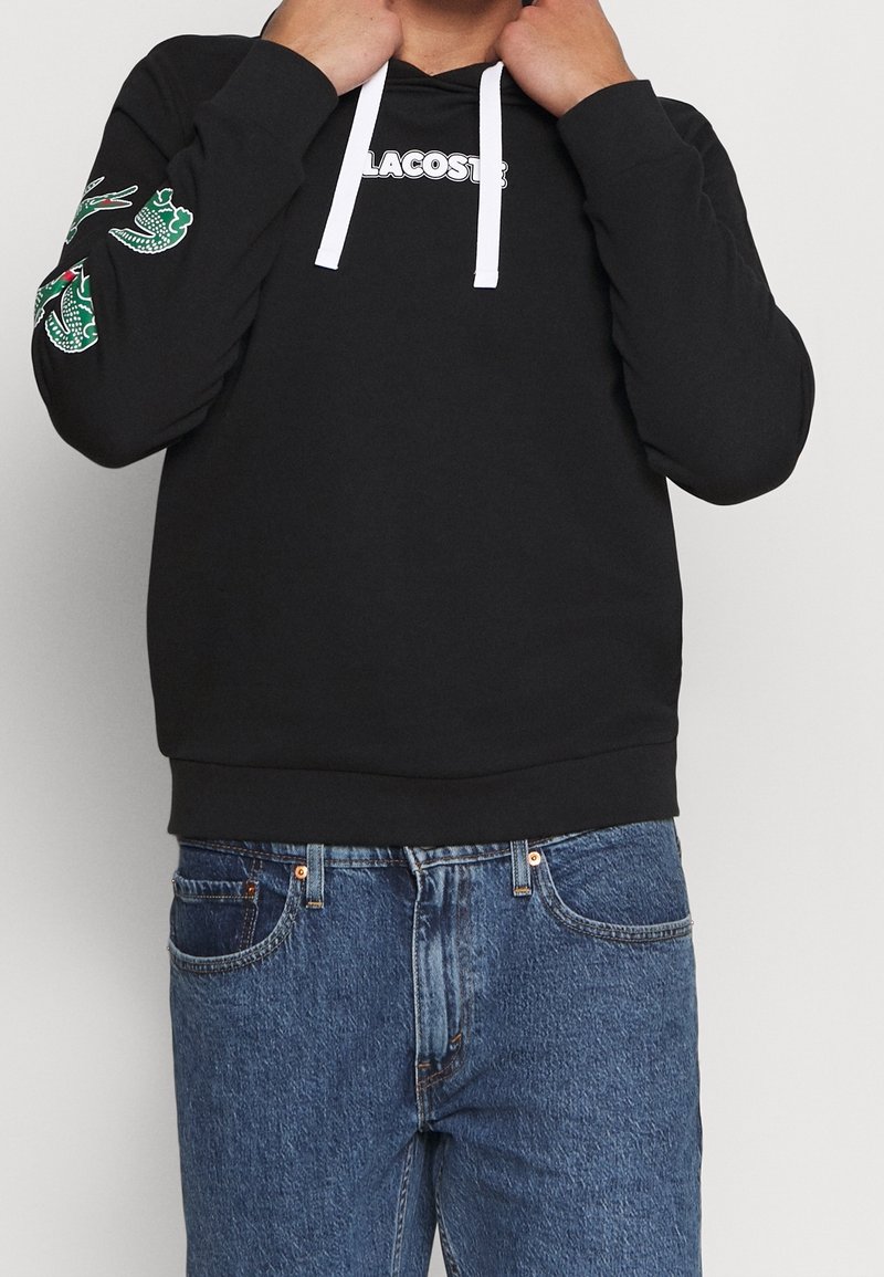 Personne portant un sweat à capuche noir Lacoste avec des cordons blancs et des logos crocodile verts sur la manche gauche et un jean bleu.