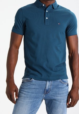 Mand iført en teal kortærmet polo med knapper og lyseblå jeans, stående med armene afslappet langs siden.