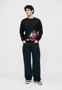 Νέος άνδρας με σγουρά μαλλιά που φοράει μαύρο φούτερ με κόκκινο και λευκό καρτούν πρόσωπο, φαρδιά μαύρη παντελόνια και ασπρόμαυρα αθλητικά παπούτσια.