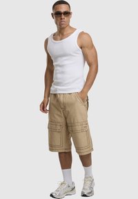 Beige Cargo-Shorts mit Cargo-Taschen und kontrastierenden Nähten, kombiniert mit einem weißen Tank-Top und silbernen Sportschuhen, accessoriert mit einer Sonnenbrille.