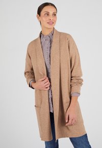 Cardigan beige con un fit morbido, apertura frontale e due tasche frontali, realizzato in materiale morbido. Indossato sopra una camicia a righe e jeans.