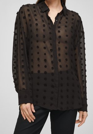 Femme portant un chemisier noir transparent à manches longues avec des pois texturés et un pantalon noir, debout avec une main reposant près de la hanche.