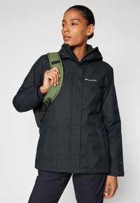 Black waterproof jacket with a hood, featuring a zip-up front, side pockets, and a logo on the chest; paired with an olive green backpack.