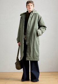 Cappotto lungo verde oliva con chiusura a zip e a pressione, dotato di cappuccio con bordo in pelliccia, due tasche laterali e un tessuto testurizzato. Abbinato a pantaloni scuri a zampa d'elefante.