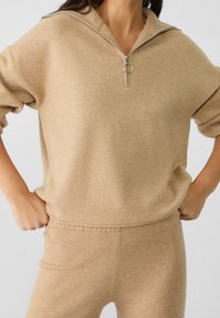 Beżowa bluza z zamkiem błyskawicznym, wykonana z miękkiego materiału, z prążkowanym kołnierzem i mankietami, z metalowym zamkiem oraz luźnym krojem.