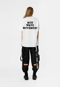 Camiseta de algodón blanca con "MOVIMIENTO MOD WAVE" impreso en negro, combinada con pantalones cargo negros que presentan múltiples bolsillos y acentos naranjas.