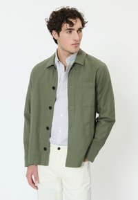 Mladý muž s kučeravými vlasmi má na sebe olivovo zelenú zapínaciu bundu, pruhované tričko a biele nohavice, pozerá sa do strany s jednou rukou v vrecku.