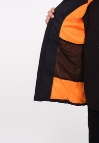Zwarte jas met oranje binnenvoering, voorzien van een mesh pocket en een gevoerde voering. De buitenkant heeft een gladde textuur en is licht glanzend.
