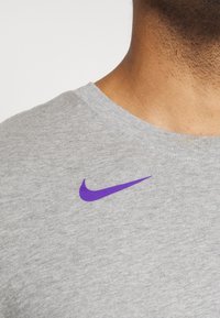 Gros plan sur le cou et l'épaule d'une personne portant une chemise gris clair avec un petit logo Nike Swoosh violet près du col.