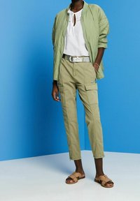 Giacca bomber trapuntata verde chiaro, blouse bianca a trama, pantaloni cargo oliva con tasche laterali, sandali piatti beige su uno sfondo blu.