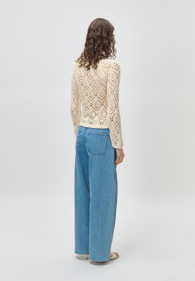 Γυναίκα με σγουρά μαλλιά φοράει κρεμ πουλόβερ πλέξης και μπλε τζιν παντελόνι με ψηλή μέση και φαρδύ πόδι, στέκεται γυρισμένη πλάτη σε απλό ανοιχτό φόντο.