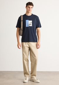 Mladý muž oblečený v námořnickém grafickém tričku, béžových kalhotách, béžových teniskách a béžové crossbody tašce stojí proti jednolitému světlému zdi.