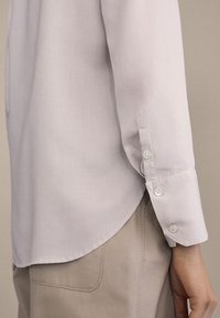 Jasnoliliowa koszula z długim rękawem, zapinana na guziki. Posiada zaokrąglony dół i białe guziki przy mankietach, wykonana jest z gładkiego materiału.