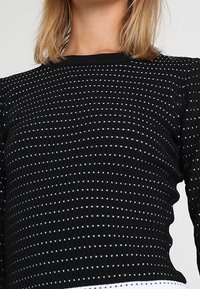 Nainen, jolla on päällä musta pitkähihainen neule, jossa on pieniä valkoisia neliökuvioita ja istuva pyöreä pääntie.