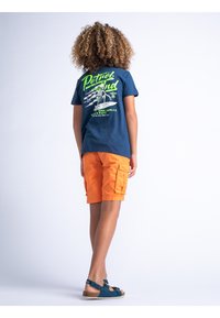 Marineblauw T-shirt met groene grafische print op de achterkant, gecombineerd met oranje cargoshorts en turquoise sandalen. Korte mouwen en casual pasvorm.
