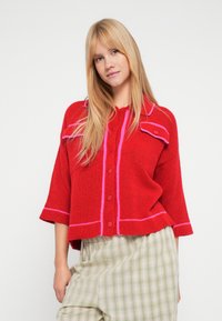 Sarkans adīts džemperis ar rozā akcentiem, garām piedurknēm, apaļu apkakli un priekšējām kabatām. Kombinēts ar zaļām rūtainām biksēm.