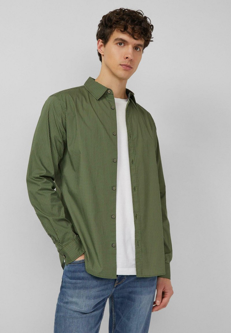 Junger Mann mit lockigem Haar, der ein grün gestreiftes Hemd über einem weißen T-Shirt und blauen Jeans trägt, steht vor einem schlichten grauen Hintergrund.