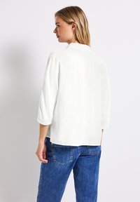 Blusa blanca de corte suelto con mangas de tres cuartos y cuello. Textura suave, dobladillo recto, combinada con jeans de mezclilla azul.