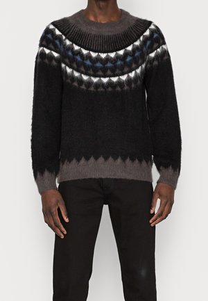 Mężczyzna w czarno-szarym swetrze z moheru z geometrycznym wzorem na klatce piersiowej i górnej części rękawów, w połączeniu z czarnymi spodniami.