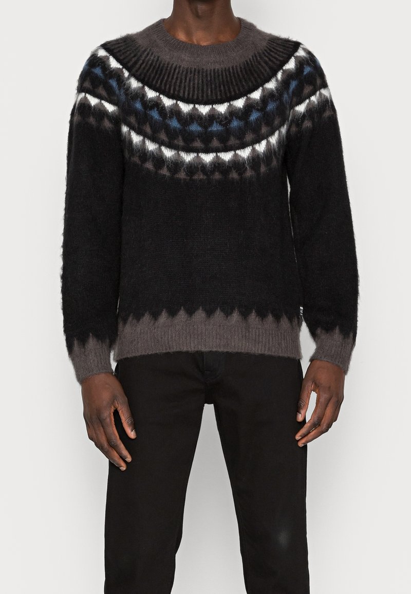 Homme portant un pull en mohair noir et gris avec un motif géométrique sur la poitrine et le haut des manches, associé à un pantalon noir.