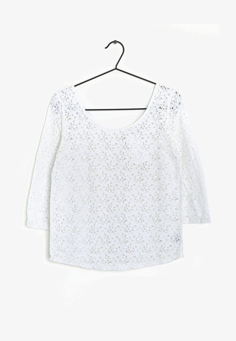 Blusa bianca a maniche lunghe in pizzo floreale con scollo rotondo appesa a una gruccia nera contro uno sfondo bianco semplice.