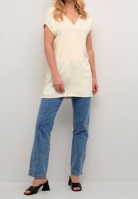 Tunique ample couleur crème avec un décolleté en V et des manches roulées, associée à un jean droit bleu clair et des sandales à talons noires.