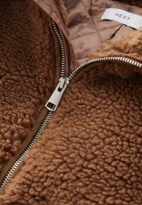 Fermeture éclair argentée partiellement ouverte sur une veste polaire texturée marron avec doublure matelassée et étiquette de vêtement visible indiquant "NEXT".