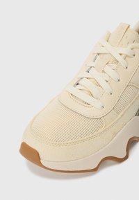 Beige sportsko med mesh-overdel og suedetekstur, med en tykk såle, flate snører og et strømlinjeformet designelement.