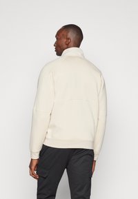 Felpa beige chiaro con colletto alto, maniche raglan e orlo a coste, abbinata a pantaloni cargo scuri, mostrata da dietro.