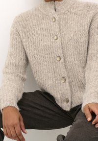 Cárdigan de punto texturizado gris claro con cuello alto y grandes botones crema, llevado sobre pantalones gris oscuro.