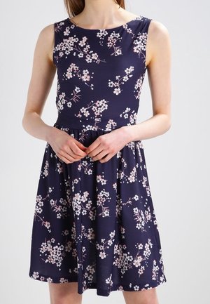 Femme portant une robe bleu marine sans manches avec un imprimé floral rose et blanc, mains jointes à la taille.
