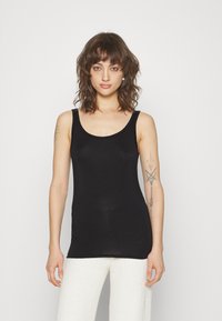 Black ribbed tank top with a scoop neckline, made of lightweight fabric. Model wearing it with white textured trousers.