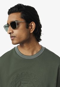 Groene sweatshirt met geborduurd logo, geribbelde kraag en grijze accenten. Slanke, hoekige zonnebril met donkere lenzen en metalen frame.