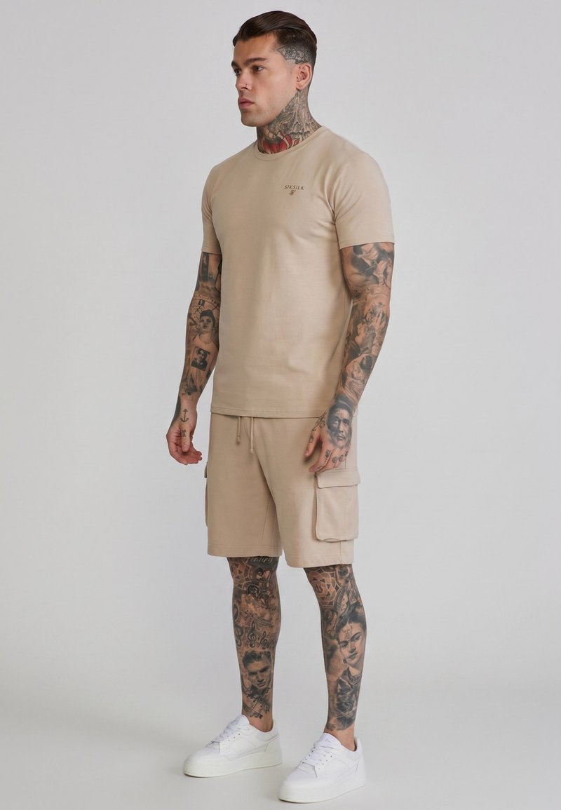 Man met tatoeages over het hele lichaam, dragend beige T-shirt en cargoshorts, staand met witte sneakers tegen een effen achtergrond.