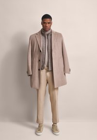Beiger Wollmantel mit großen Revers, Knopfverschluss; grauer Rollkragenpullover darunter; cremefarbene Hose; beige Sneaker. Schlicher Hintergrund.
