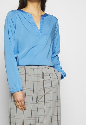 Woman wearing a long-sleeve light blue blouse tucked into high-waisted beige pants with a plaid pattern and vertical dark stripes.