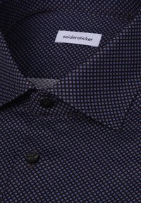 Chemise sombre avec petits motifs carrés bleus, boutons noirs et étiquette blanche à l’intérieur du col portant la mention "seidensticker".