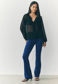 Must mustriga pluus volangidega, mida on sobitatud kitsaste siniste kellukeste pükstega. Mudel kannab musti sandaale, luues juhusliku kuid samas elegantsse välimuse.