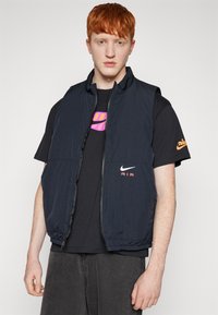 Μαύρο νάιλον γιλέκο με κλείσιμο φερμουάρ και ψηλό γιακά. Διαθέτει δύο μπροστινές τσέπες και το λογότυπο της Nike με την ένδειξη "AIR" σε κόκκινο χρώμα στο στήθος.