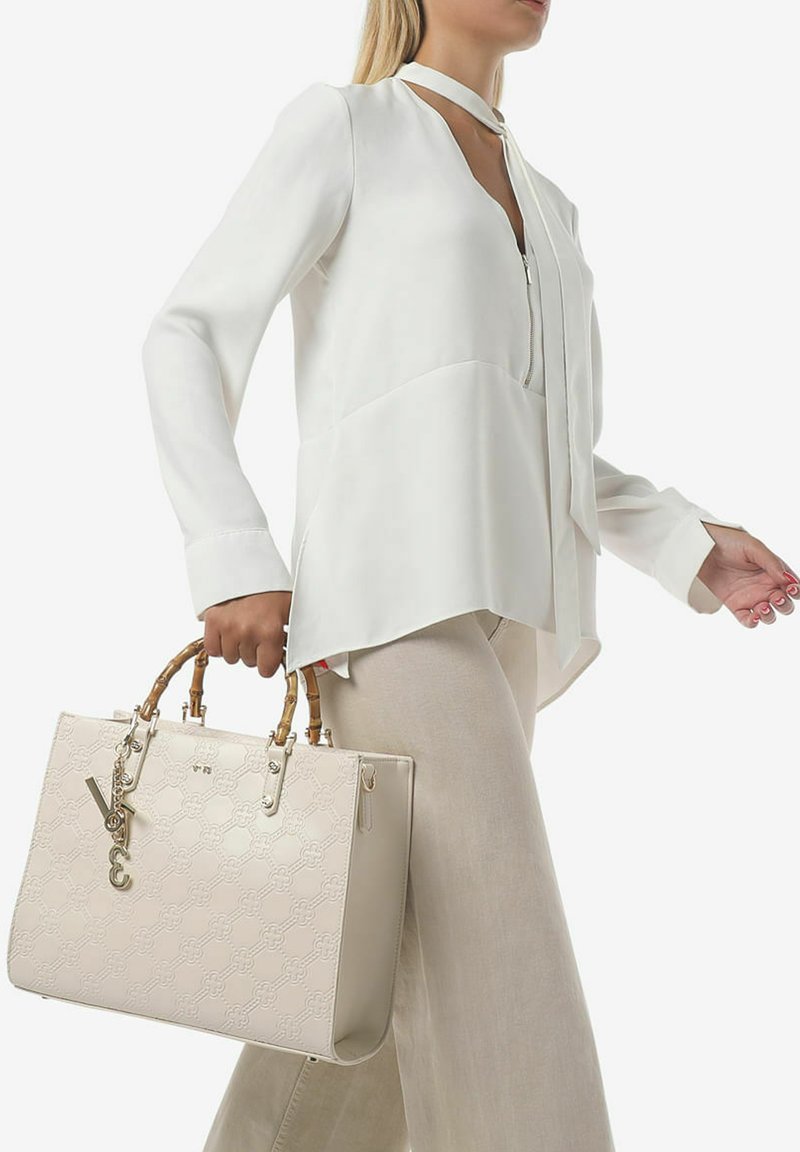 Sac à main beige avec motif gaufré, poignées en bambou et accents en métal. Le sac a une forme rectangulaire et est porté avec une blouse légère.