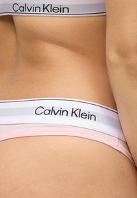 Τα εσώρουχα Calvin Klein διαθέτουν μια απαλό ροζ υφασμάτινη λωρίδα με λευκή ελαστική μέση που περιλαμβάνει λογότυπο και γκρι διακοσμητικές ρίγες.
