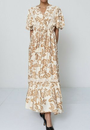 Robe maxi à motifs floraux en beige et crème, avec des manches courtes, une taille cintrée et une bordure en dentelle à l'ourlet. Portée avec des talons noirs.