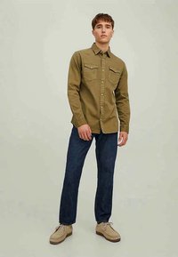 Camicia verde oliva Button-up con due tasche sul petto, abbinata a jeans blu scuro e scarpe beige, con una vestibilità rilassata e colletto.