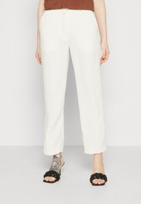 Pantaloni bianchi a gamba larga con una texture liscia, caratterizzati da un davanti piatto e passanti per cintura. Il modello indossa sandali neri intrecciati.