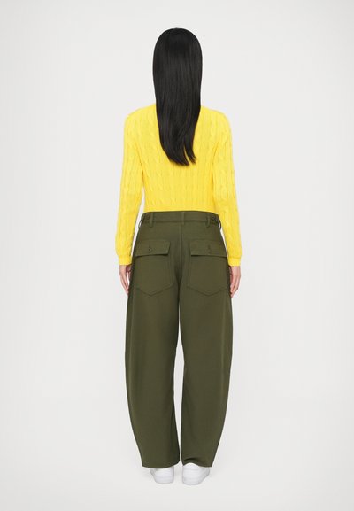 Κίτρινο πουλόβερ με πλέξη καλσόν, πράσινο παντελόνι φαρδύ με μεγάλες πίσω τσέπες και άσπρα αθλητικά παπούτσια, βλέποντας από πίσω πάνω σε λευκό φόντο.