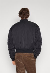 Schwarze Bomberjacke mit gerafften Ärmeln, geripptem Kragen und Bündchen, glatte Textur. Kombiniert mit braunen Cordhosen, von hinten betrachtet.