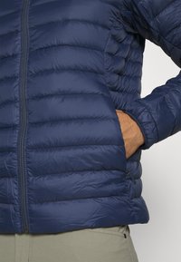 Mornarčasto plava puf jakna s kvadratnim uzorkom, prednji patentni zatvarač i bočne džepove. Čini se da je materijal lagan i vodootporan.