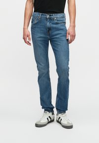 Pessoa vestindo jeans justos azuis e ténis brancos com riscas pretas, de pé contra um fundo claro e liso.